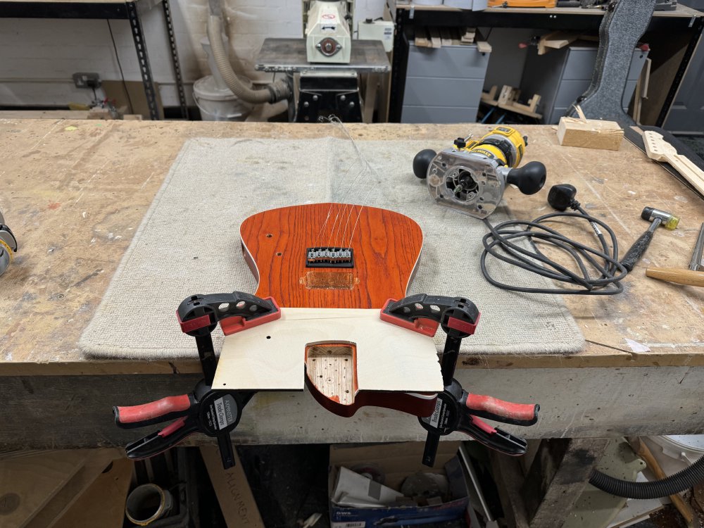 A photo of the guitar body clamped to a workbench, with a protective plywood jig over most of it, leaving just the neck pocket exposed. Next to it sits a palm-router.