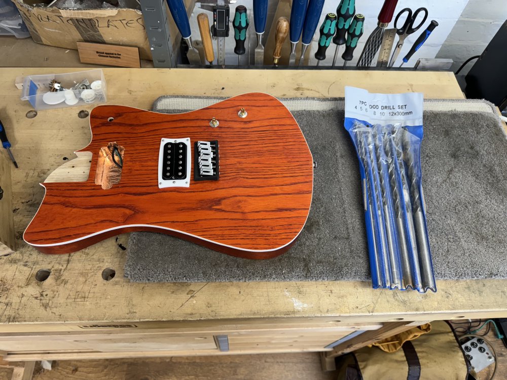 A photo of the guitar body on the workbench, without its neck again, and one of the pickups missing. Next to the body sits a pack of foot-long drill bits.
