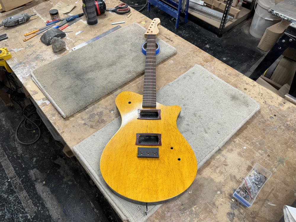 A photo of a work in progress guitar on the workbench, with both the body and neck in place, but missing pickups, tuners, etc. but with the pickup mount rings and bridge placed in position.