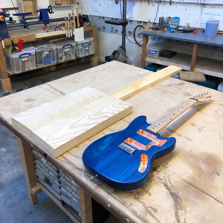 The workshop workbench, on which sits some untouched blocks of woods arranged in the position of the neck and body of a guitar, and then a late stage guitar build next to it.