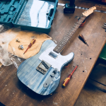 A denim blue t-style guitar sits on the workbench, with a pearloid white pickguard sporting a chrome-capped humbucker. No strings or controls yet added, this is an in-progress shot.