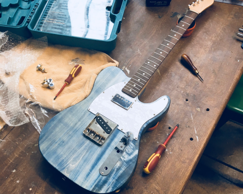 A denim blue t-style guitar sits on the workbench, with a pearloid white pickguard sporting a chrome-capped humbucker. No strings or controls yet added, this is an in-progress shot.