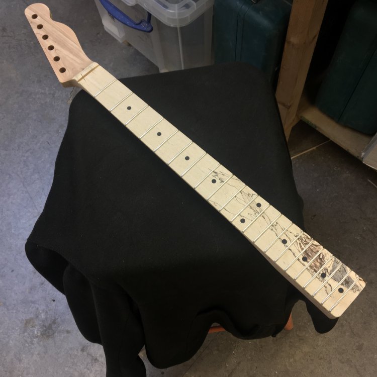 A work in progress guitar neck sits on a cloth covered stool in the workbench. The neck is shaped and has frets in, and the fretboard has a nice spalted wood look, where black marks in the light wood look a little like flames at the bottom of the neck.