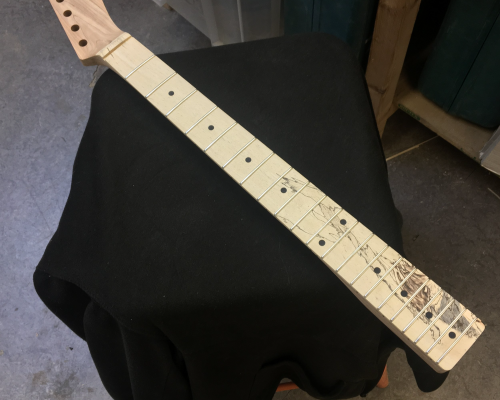 A work in progress guitar neck sits on a cloth covered stool in the workbench. The neck is shaped and has frets in, and the fretboard has a nice spalted wood look, where black marks in the light wood look a little like flames at the bottom of the neck.