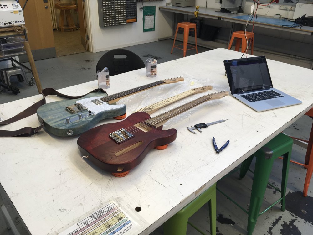 Two t-style electric guitars sit on a workbench, a blue one that is finished, and a red one that is clearly work in progress as it has no pickups etc. In between them sits a third neck of a similar shape.