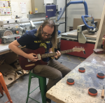 A photo of me sat in the workshop testing a work in progress electric guitar that's missing the electronics but is otherwise strung up.