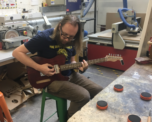 A photo of me sat in the workshop testing a work in progress electric guitar that's missing the electronics but is otherwise strung up.