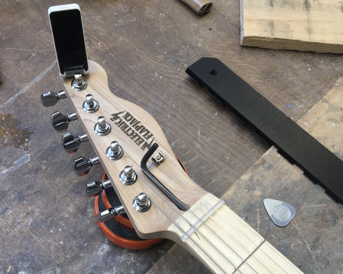 A close up of the headstock of a guitar neck, strings on, with clip on tuner and allen key in the truss-rod socket.