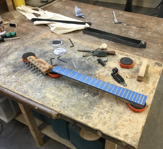 A finished guitar neck sits on the workbench surrounded by tools. The fretboard has been covered in masking tape leaving only the frets clear.