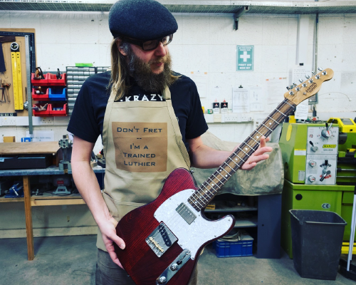A photo of me in the workshop holding a finished guitar wearing an apron that has printed upon it 'Don't Fret - I'm a Trained Luthier'