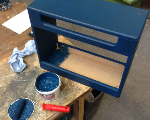 A empty amp cabinet sits on the workbench being painted with a blue substance. You can see a small roller and tub of paint beside it.