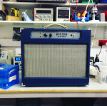 A blue all-in-one amp sits on a workbench. The main front face is taken up with silver grill fabric covering the speaker, and there is a silvery control panel with all the controls at the top.