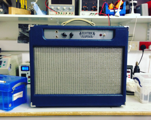 A blue all-in-one amp sits on a workbench. The main front face is taken up with silver grill fabric covering the speaker, and there is a silvery control panel with all the controls at the top.