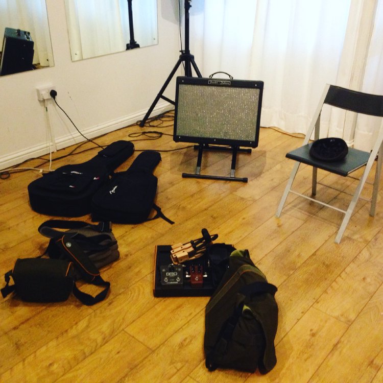 A studio space with a Fender Deluxe amplifier on a stand, a couple of guitar bags, misc cables, and a chair.