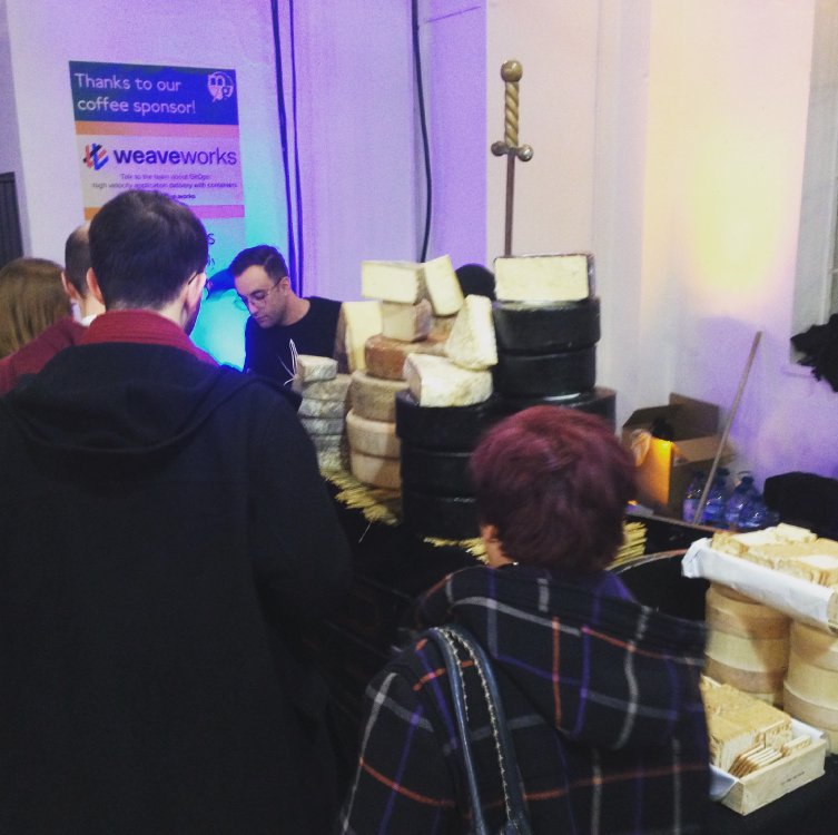 A picture of a stand with a queue of people at it. On the stand are several mounds of cheese wheels stacked 5 ot 6 high.