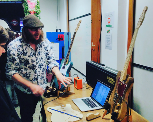 A photo of me stood by a table with guitars and a laptop on, explaining to someone how guitars are designed and built.
