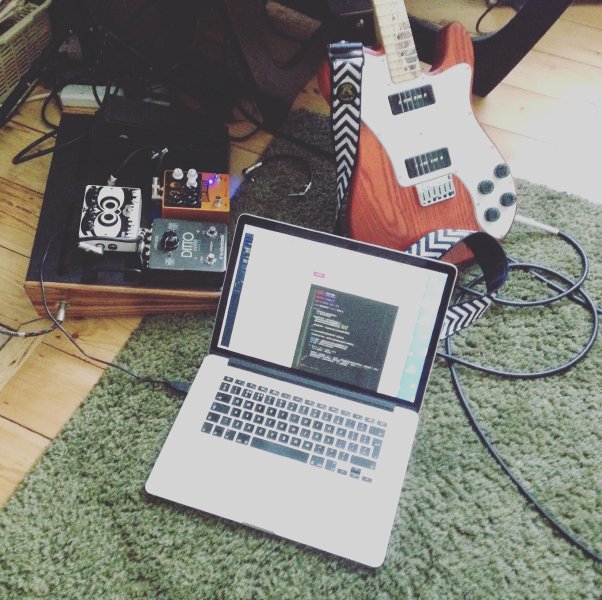 A photo of a guitar pedalboard with a laptop next to it showing code on the screen.