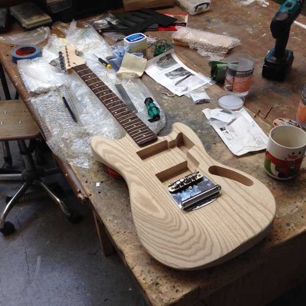 A work in progress guitar sits on the workbench. The body is shaped and has cavities cut out, and has a shiny bridge attached to it, but is otherwise unfinished.