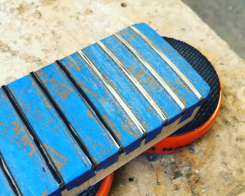 A guitar neck sits on the workbench, with the fretboard covered in masking tape to leave just the ferts showing. Half the frets have black sharpie on them, waiting to be shaped, the rest are shiny where they've been worked on.