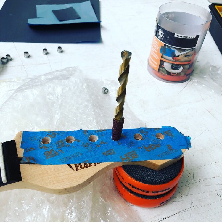 The headstock of a guitar, yet to have tuners mounted. The face of the headstock is covered in masking tape, and in one of the tuner holes is a drill bit with sand paper wrapped around it.