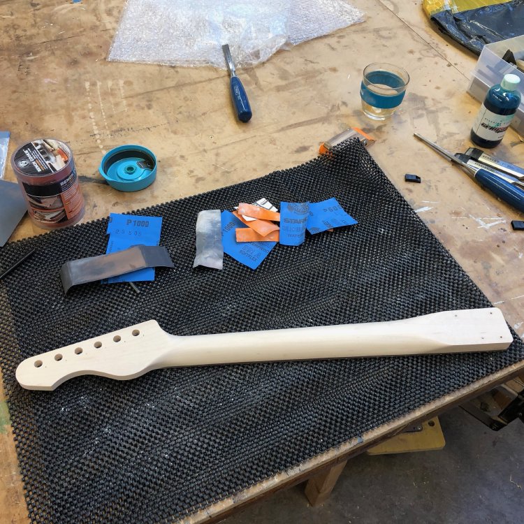 A guitar neck sits on the workbench, rear side up, and is surrounded by bits of sandpaper.