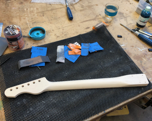 A guitar neck sits on the workbench, rear side up, and is surrounded by bits of sandpaper.