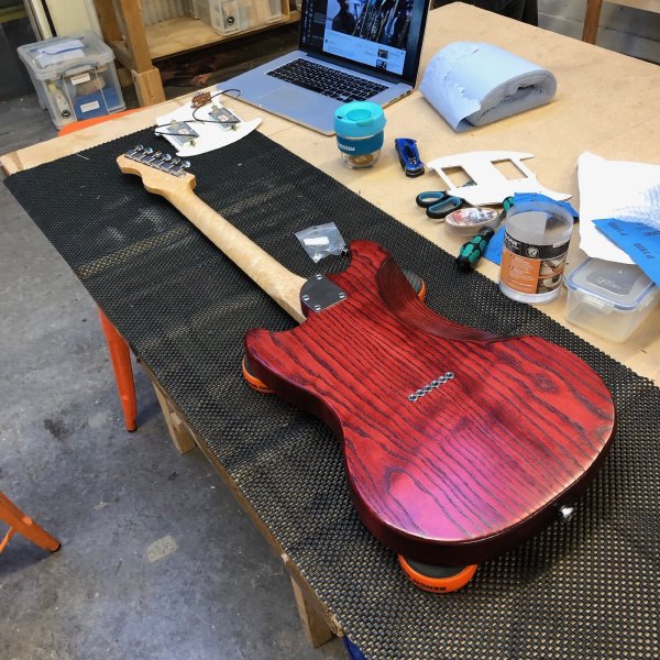 A mustang-style guitar sits on the workbench, rear-side up. It has a crimson body and you can see six string ferrules in the back of it, and it has a curved neck-pocket. The neck is maple with vintage style tuners.