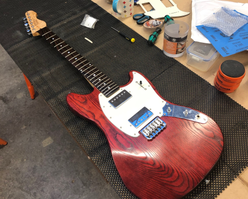 A mustang-style guitar sits on the workbench. It has a crimson body and a white pickguard, into which are installed two chrome covered humbuckers. The neck is maple with a wenge fretboard.