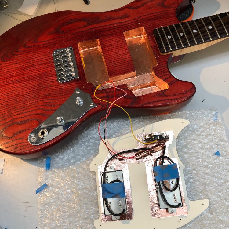 A close up of the guitar body on the workbench, with the pickguard off to the side. The body cavities are lined with copper tape, and you can see some wires going over to the pickguard in which is mounted the pickups and a selector switch.