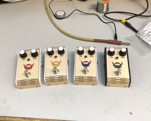 A row of four guitar effects pedals sits on the workbench. Each one has a laser-etched wooden face with a beard logo, each pedal having the beard stained a different colour.