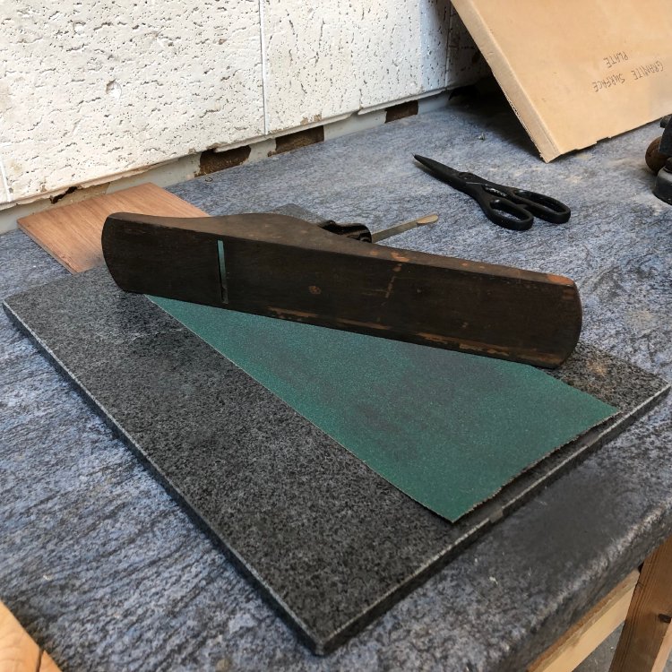 An old hand-plane sits on its side upon a sheet of sandpaper that has been placed on a granite block that has a known true surface.