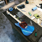 A striking blue electric guitar with a red tort pickguard and maple neck sits on the workbench as it undergoes final tweaking.