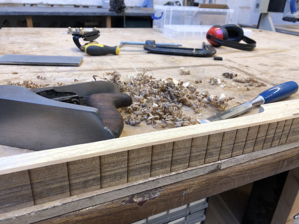 A close up of the neck after being worked on, showing the seam between fretboard and neck is completely level and smooth. Behind it you can see a lot of wood shavings, a hand plane, and a chisel.