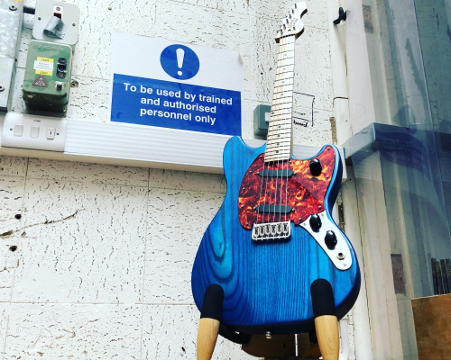 A blue mustang style guitar with a marbled red pickguard and a maple nect sits in a stand in the workshop, next to a workshop sign that reads 'To be used by trained and authorized personnel only.'