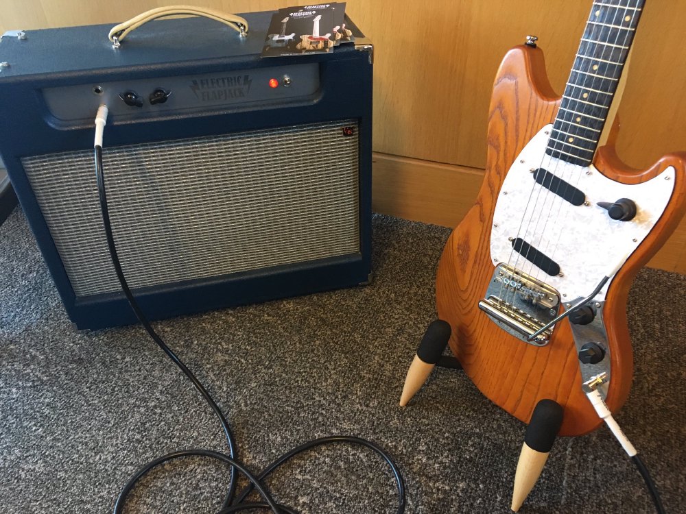 An orange coloured guitar sits on a stand next to a blue coloured amplifier, with a cable running between them.