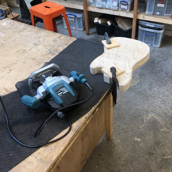 A block of wood that has been cut to roughly the shape of a guitar on the bandsaw sits clamped to a workbench, with an acrylic template of the final shape stuck to it. Beside it sits a plunge router.