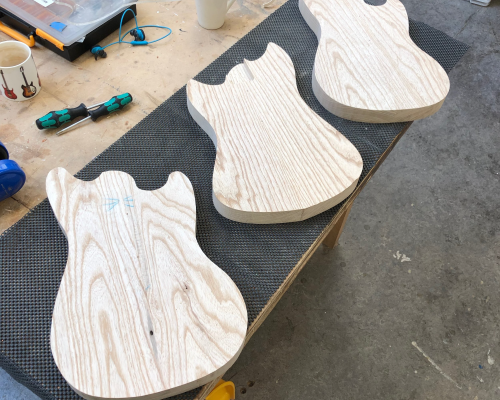 Three early stage guitar bodies, all just cut out as profiles from blocks of wood, sit on the workbench in a row.