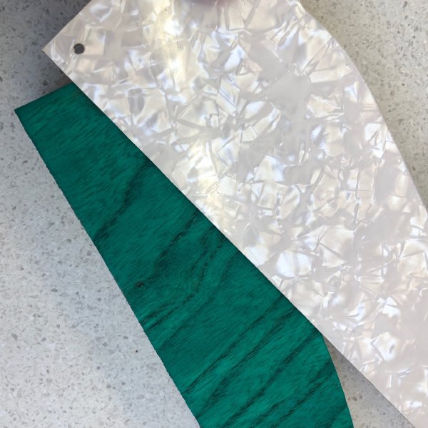 A photo of an offcut of swamp-ash stained green, and over it an offcut of white pearloid pick-guard material.