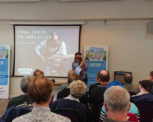 I am presenting to a crowded room, showing the audience somethinb about the guitar I'm holding, in front of a slide titled 'From Frets to Amplifiers'
