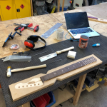 A messy workbench shows a guitar neck in late build process, with about a third of the frets inserted. Beside the neck sits a hammer with plastic ends, a fret-slot saw, a laptop, some fret-wire, and misc other tools.