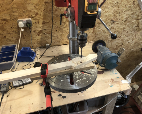 A roughly shaped guitar neck is on a pillar drill, with the holes for the tuning pegs being drilled.