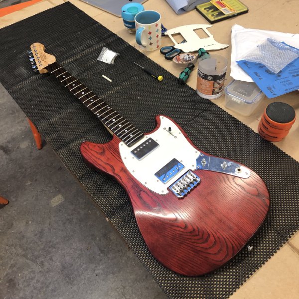 A cherry-red mustang-style guitar sits upon a workbench, looking almost complete. On the face of the body is a white plastic pick-guard into which two chrome-covered humbucker pickups are mounted.