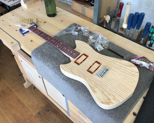 An in-progress pointy-offset style guitar build sits on the workbench surrounded by bags of parts. The neck, which maple with a purpleheart fretboard, is mostly completed but un-oiled, and the body is shaped and sanded, but is yet to have holes put in it for pickups and controls. On top of the body sit a bunch of parts placed to give an idea of how it'll look when finished.