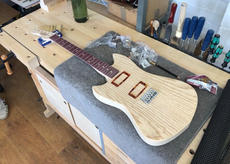 An in-progress pointy-offset style guitar build sits on the workbench surrounded by bags of parts. The neck, which maple with a purpleheart fretboard, is mostly completed but un-oiled, and the body is shaped and sanded, but is yet to have holes put in it for pickups and controls. On top of the body sit a bunch of parts placed to give an idea of how it'll look when finished. To one side is a half eaten snack bar.