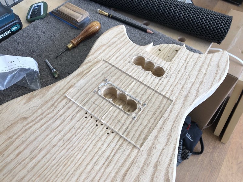 The guitar body is now back on the workbench, with three interlinked holes that have removed most of the material for the two pickup cavities. Over one of the cavities is mounted a laser-cut acrylic template for routing out the rest of the material.