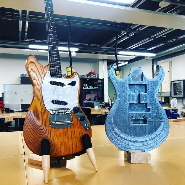 On a workbench in a large room sits a custom guitar I made with an orange-stained ash body and maple neck with wenge fretboard, beside which sits a full sized 3D-print of a guitar body.