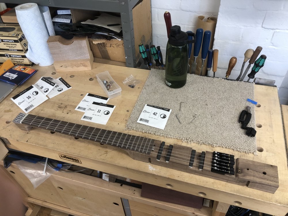 The wooden core of my älgen guitar, which runs the neck and center of body, sits on a workbench. It has some strings on it, and is surrounded by string packets.