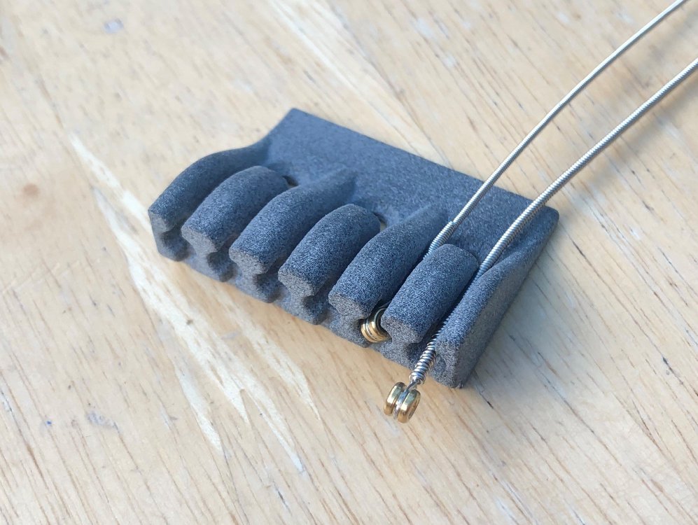 A photo of a 3D-printed plastic part that has 6 channels in it that end in little thimbles. In one channel sits a guitar string with the ball end sat in the thimble. In another channel a string sits over the channel as it's too wide to fit in.