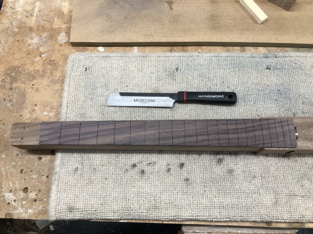 A photo of a top down view of the neck of a guitar build, showing the fretboard with the fret slots cut, and next to the neck is a small saw japanese pull saw.