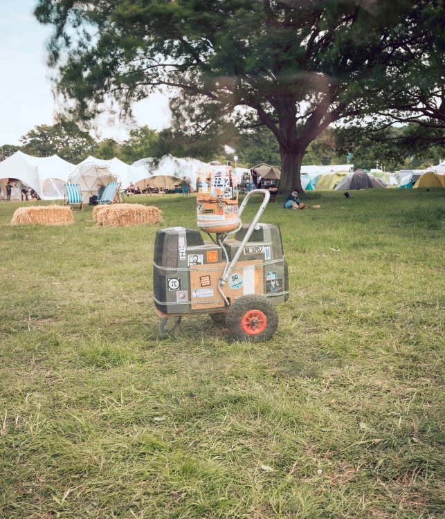 A photo of a simple robot contraption that looks a bit like a suitcase with wheels and a handle with a propane tank on top. 